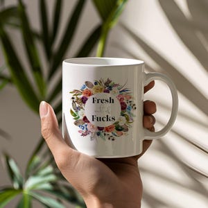 May include: White ceramic mug with a floral wreath design and the text "Fresh out of Fucks". The wreath features colorful flowers and leaves. The mug has a large handle and is held by a hand.