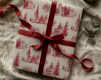 May include: A present wrapped in paper with a red toile pattern of buildings and trees, tied with a red satin ribbon and bow. The gift is on a white surface, with a candle and greenery in the background.
