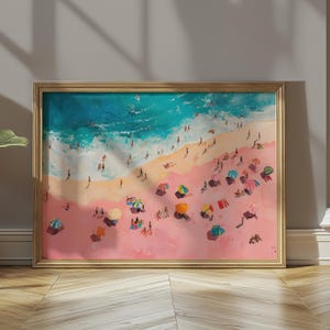May include: A framed beach scene painting with a gold-colored frame. The artwork depicts a pink sandy beach with people, colorful umbrellas, and turquoise ocean waves. The painting is displayed on a wooden floor.