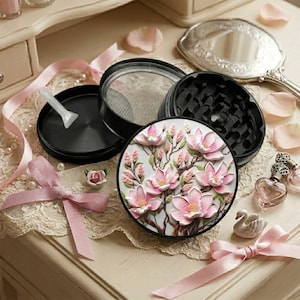 May include: A black circular herb grinder featuring a decorative lid with a raised, detailed illustration of pink magnolia flowers on branches. The set includes a matching black base, a mesh screen insert, and a small white plastic scraping tool.