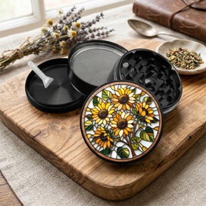 May include: A disassembled black herb grinder with a sunflower design. The grinder features a stained glass-style sunflower pattern on the top, with yellow, orange, and green colors. A small white scoop is included.