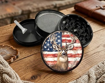 May include: A black, four-piece herb grinder with a deer and American flag design. The grinder is round and has a mesh screen and a small plastic scraper. The design features a buck with antlers set against a backdrop of the American flag.