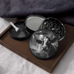 May include: Black and white grinder with a cow and UFO design. The grinder is disassembled, showing the different parts, including a screen and a small scoop. The cow graphic features a surprised expression under a UFO.