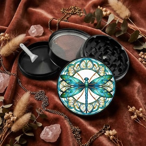 May include: A black metal herb grinder featuring a circular top lid with a vibrant blue and teal dragonfly graphic in a stained glass style. The grinder is disassembled into three parts, revealing a mesh screen, grinding teeth, and a small white scraper.