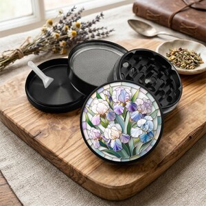 May include: Black, multi-piece herb grinder with a floral iris design. The grinder is disassembled, showing the various components, including a mesh screen and a small plastic scoop. The design features white, purple, and blue irises.