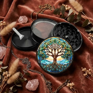 May include: A black metal herb grinder featuring a circular top lid with a stained glass style design of a tree with blue and green leaves and exposed roots. The grinder is disassembled into three parts, including a small white plastic scraper.
