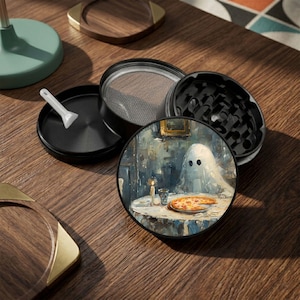 May include: A multi-part black metal herb grinder featuring a circular lid with an artistic print of a ghost sitting at a table with a pizza. The set includes a mesh screen component, a bottom collection chamber, and a small white plastic scraper tool.