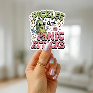 May include: A sticker with the text "Pickles and Panic Attacks" and a cartoon pickle wearing sunglasses. The sticker has a white border and is held by a hand with gold rings and iridescent nails. The design includes flowers and lightning bolts.