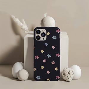 May include: A black phone case adorned with a repeating pattern of small, colorful flowers in pink, yellow, and blue. The case is shown upright, with the camera lenses visible. The design is simple and floral.