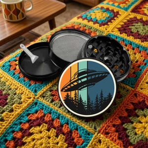 May include: A black herb grinder with a screen and a small scoop, next to a round sticker with a retro design of a UFO over a forest. The items are on a colorful, crocheted blanket.