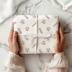 May include: A wrapped gift with a white background featuring a pattern of brown paw prints and dog bones. The gift is tied with a white ribbon. The gift is held by a person wearing a white long-sleeved shirt.
