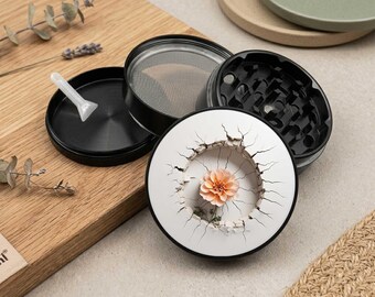 May include: A black multi-piece herb grinder with a white top lid featuring a cracked circular design and a central peach-colored flower. The set includes a fine mesh screen, a black base component, and a small white plastic scraper tool.