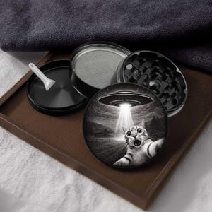 May include: A black and silver herb grinder with a cat and UFO graphic. The grinder is disassembled, showing the different parts, including a screen and a scoop. The cat graphic depicts a surprised cat being beamed up by a UFO.