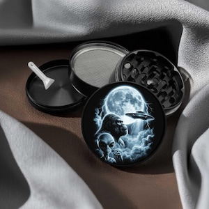 May include: A black multi-piece herb grinder featuring a circular lid with a graphic print of an alien, a gorilla, a full moon, lightning, and a flying saucer. The set includes a metal mesh screen, a grinding chamber, and a small white plastic scraper.