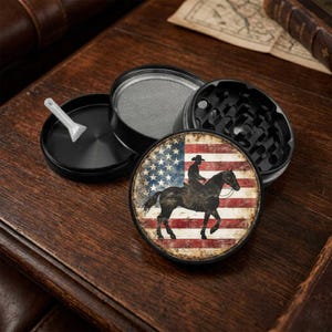 May include: A black and silver grinder with a cowboy riding a horse graphic over an American flag design. The grinder is disassembled, showing the different components, including a sifter and a small scoop.