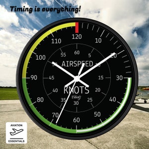 May include: Aviation-themed wall clock with a black face, white hands, and a black frame. The clock face displays airspeed in knots, with the text "Timing is everything!" at the top. The clock has a green and yellow arc and the words "Aviation Essentials".