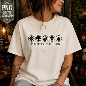 May include: A cream-colored short-sleeve t-shirt featuring a black graphic print of five circular symbols representing a sun, tree, wave, skull, and water drop, with the text "MAGIC IS IN THE AIR" printed in black lettering below the icons.