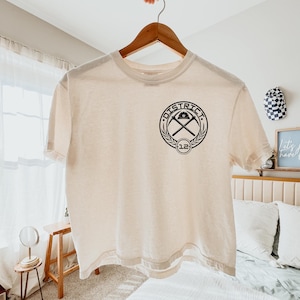 K&ouml;nnte beinhalten: Cremefarbenes T-Shirt mit schwarzem Aufdruck. Das kreisf&ouml;rmige Design zeigt das Wort "DISTRICT", gekreuzte Spitzhacken und die Zahl "12". Das T-Shirt h&auml;ngt an einem Holzb&uuml;gel.