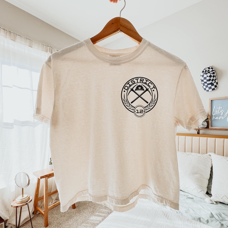 K&ouml;nnte beinhalten: Cremefarbenes T-Shirt mit schwarzem Aufdruck. Das kreisf&ouml;rmige Design zeigt das Wort "DISTRICT", gekreuzte Spitzhacken und die Zahl "12". Das T-Shirt h&auml;ngt an einem Holzb&uuml;gel.