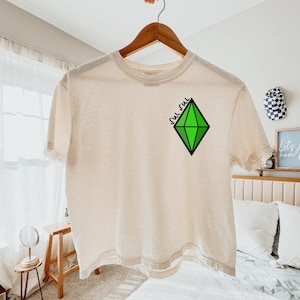 以下が含まれることがあります： グリーンのダイヤモンドグラフィックと「Sul Sul」の文字が入ったクリーム色のTシャツ。グラフィックは、緑色の塗りつぶしと黒いアウトラインを持つ様式化されたダイヤモンドの形です。Tシャツは木製のハンガーに吊るされています。
