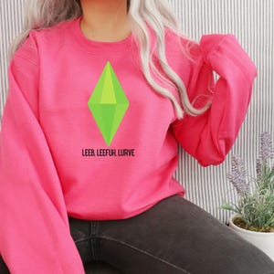 May include: A bright pink sweatshirt featuring a large green diamond graphic. Below the diamond, the text reads "LEEB, LEEFUH, LURVE". The sweatshirt has long sleeves and a crew neck.