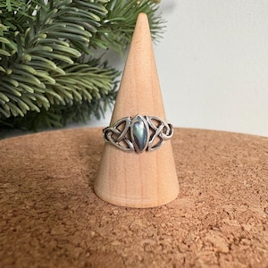 Puede incluir: Un anillo plateado con una piedra central ovalada e iridiscente, engastada en un diseño de nudo celta. El anillo se exhibe sobre un soporte de madera en forma de cono, mostrando sus intrincados detalles. Una joya.
