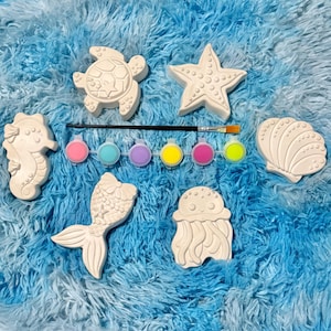 May include: A set of six white plaster sea-themed figurines including a turtle, starfish, seahorse, seashell, mermaid tail, and jellyfish, accompanied by a small paintbrush and a strip of six small paint pots in pink, blue, purple, yellow, and green.