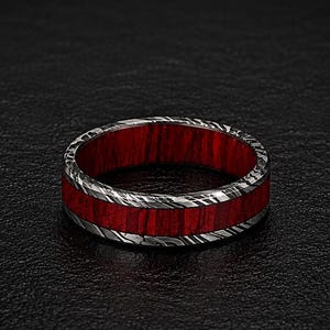 May include: A ring featuring a central band of vibrant red material inlaid between two outer edges of dark, textured metal with a swirling, marbled pattern. The design is symmetrical, showcasing a contrast between the bright red center and the dark, metallic, patterned exterior.