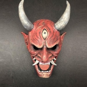 Può includere: Una maschera giapponese Oni rossa e bianca con un terzo occhio e denti aguzzi. La maschera ha un'espressione feroce ed è un simbolo tradizionale di forza e potere.