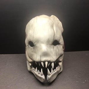May include: A white, detailed, and textured mask with a wide, toothy grin. The mask has a cracked and weathered appearance, suggesting age and use.