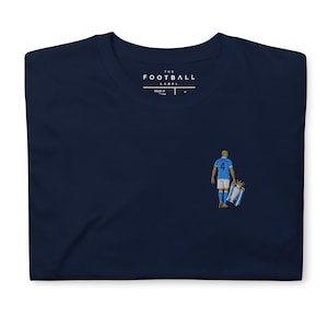 以下が含まれることがあります： ネイビーブルーのTシャツ。右側に背番号4のライトブルーのユニフォームを着てトロフィーを運ぶサッカー選手の小さな刺繍グラフィックが施されています。首元の内側のラベルには「THE FOOTBALL LABEL」というテキストが表示されています。