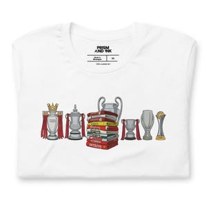 Peut inclure: T-shirt blanc avec un graphisme de trophées de football, dont le trophée de la Premier League et une coupe d'argent. Le design comprend également une pile de livres et d'autres récompenses. Le t-shirt porte le texte "PRISM AND INK" sur l'étiquette.