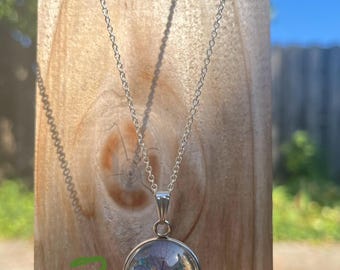 May include: A silver-toned necklace with a round pendant. The pendant encases a dried blue and purple flower. The necklace hangs against a wooden background, with the number "2" written in green on the wood.