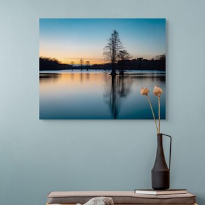 Può includere: Fotografia incorniciata di un paesaggio lacustre al tramonto, con alberi riflessi nell'acqua. Sotto la fotografia c'è una panca di legno con un cuscino grigio e un cassetto. Un vaso nero con fiori secchi e libri sono sulla panca.