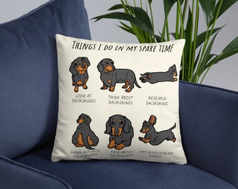 Puede incluir: Una almohada blanca con una ilustración de un dachshund negro y marrón. La almohada presenta el texto "Things I do in my spare time" y enumera actividades relacionadas con los dachshunds, como "mirar dachshunds", "pensar en dachshunds" y "ver videos de dachshunds".