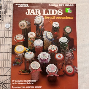 May include: A Leisure Arts craft leaflet titled Jar Lids for all occasions, featuring 17 cross-stitch designs for jar lids. The cover displays various glass jars filled with items, each topped with a decorated lid, next to a 14-inch ruler.
