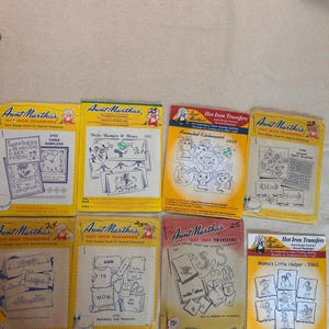 May include: Eight Aunt Martha's hot iron transfer packets featuring various embroidery designs. The yellow, white, and red paper envelopes display illustrations of samplers, kitchenware, quilt blocks, alphabets, and children, with model numbers.