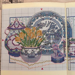 May include: A cross-stitch pattern printed on a grid featuring a blue and white ceramic bowl filled with yellow flowers, a decorative plate with a pagoda scene, and a small figurine. A Lip Edge ruler is visible at the top with inch markings.