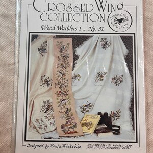 May include: A cross-stitch pattern booklet titled Crossed Wing Collection Wood Warblers I No. 31. The cover features a photograph of embroidered fabric with bird motifs, a vertical sampler strip, two small books, and binoculars in a plastic sleeve.