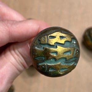 May include: A round metal cabinet knob featuring a raised gold-toned design of three stacked fish against a recessed, oxidized green background. The circular knob has a weathered, aged patina finish with visible texture across the metal surface.