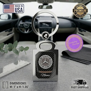 May include: A silver-toned metal keychain featuring a circular ring and a rectangular pendant with a carbon fiber texture. The pendant displays the Mercedes-Benz logo and the word Name. A ruler shows dimensions of 1 inch by 1.35 inches.