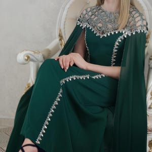 Può includere: Un abito lungo verde scuro caratterizzato da uno scollo trasparente con ricami argentati, un mantello sovrapposto e bordi decorati con perline argentate lungo il corpetto e la gonna. L'abito è abbinato a tacchi a piattaforma blu scuro con cinturini.