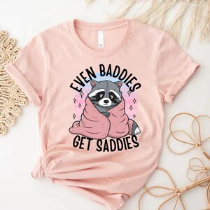 Pode incluir: Uma camiseta rosa com um gr&aacute;fico de um guaxinim enrolado em um cobertor rosa. O texto na camiseta diz "Even Baddies Get Saddies".