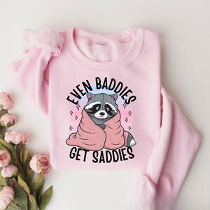 Pode incluir: Camisola rosa com um gr&aacute;fico de um guaxinim envolto em um cobertor rosa e o texto "Even Baddies Get Saddies".