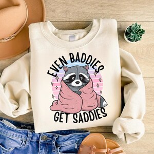 Pode incluir: Uma camisa de moletom cor creme com um gr&aacute;fico de um guaxinim enrolado em um cobertor rosa. O texto "Even Baddies Get Saddies" est&aacute; impresso em preto na camisa de moletom.