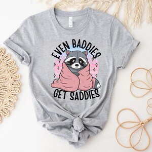 Pode incluir: Camiseta cinza com um desenho animado de um guaxinim envolto em um cobertor rosa e o texto "Even Baddies Get Saddies".