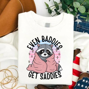 Pode incluir: Camisola branca com um desenho animado de um guaxinim envolto em um cobertor rosa e o texto "Even Baddies Get Saddies".
