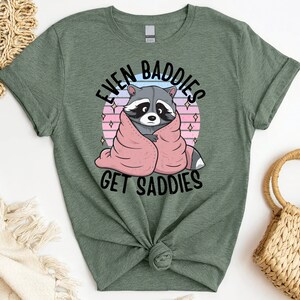 Pode incluir: Camiseta verde com um guaxinim envolto em um cobertor rosa e o texto "Even Baddies Get Saddies".