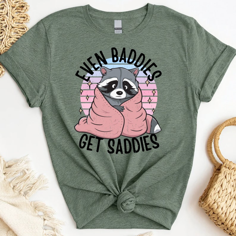 Pode incluir: Camiseta verde com um guaxinim envolto em um cobertor rosa e o texto "Even Baddies Get Saddies".