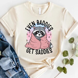 Pode incluir: Uma camiseta bege com um gr&aacute;fico de um guaxinim enrolado em um cobertor rosa. O texto "Even Baddies Get Saddies" est&aacute; impresso na camiseta.